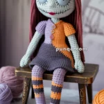 Sally from The Nightmare Before Christmas in yarn form. A crocheted doll with large stitched eyes, long coral hair, and a patchwork dress in orange and purple, seated on a wooden stool. There are balls of yarn and crochet hooks on the table in the foreground.