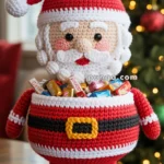 A crocheted Santa Claus container filled with assorted candies, placed on a wooden table with a blurred Christmas tree in the background.
