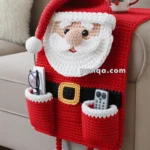 A crocheted Santa-themed armchair organizer with a Santa face and hat, featuring pockets holding a remote and eyeglasses. It hangs over the arm of a beige sofa, accented with a decorative holiday mug on the armrest.