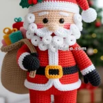 A crocheted Santa Claus doll with a red suit, white beard, and a sack of gifts over his shoulder, standing against a blurred background with a Christmas tree.
