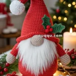 A crocheted gnome with a red hat adorned with holly leaves and berries, featuring a long white beard, is standing on a table decorated with lights and small crocheted gifts. A blurred Christmas tree is in the background.