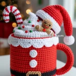 A crocheted red mug designed to resemble Santa's outfit, complete with a black belt and white buttons, topped with white whipped cream, colorful sprinkles, a candy cane, and a small gingerbread figure, all crocheted. The mug has a red handle and a Santa hat hanging off the side. The background features blurred Christmas lights and decorations.