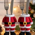 Two pairs of forks and knives dressed in knitted Santa Claus suits placed upright on a table, surrounded by Christmas decorations and lights.