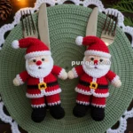 Two crocheted Santa Claus figures holding cutlery on a round, knit green placemat, decorated with white lace edges, surrounded by pine branches, pinecones, and string lights.