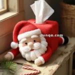 A crocheted Santa Claus tissue box cover with a cute face and white beard, placed on a wooden table next to two candy canes and pine branches, with sunlight coming through a nearby window.