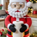 Crocheted Santa Claus figure holding a roll of paper towels, set in a kitchen with holiday decorations including string lights and Christmas ornaments.