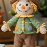 A crocheted scarecrow doll with a brown hat, a green jacket, and brown pants, featuring straw details on the hands and feet and a cheerful facial expression.