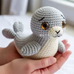 A person holds a small, crocheted seal toy with large amber eyes, featuring gray and cream-colored yarn, on a bed near a window.