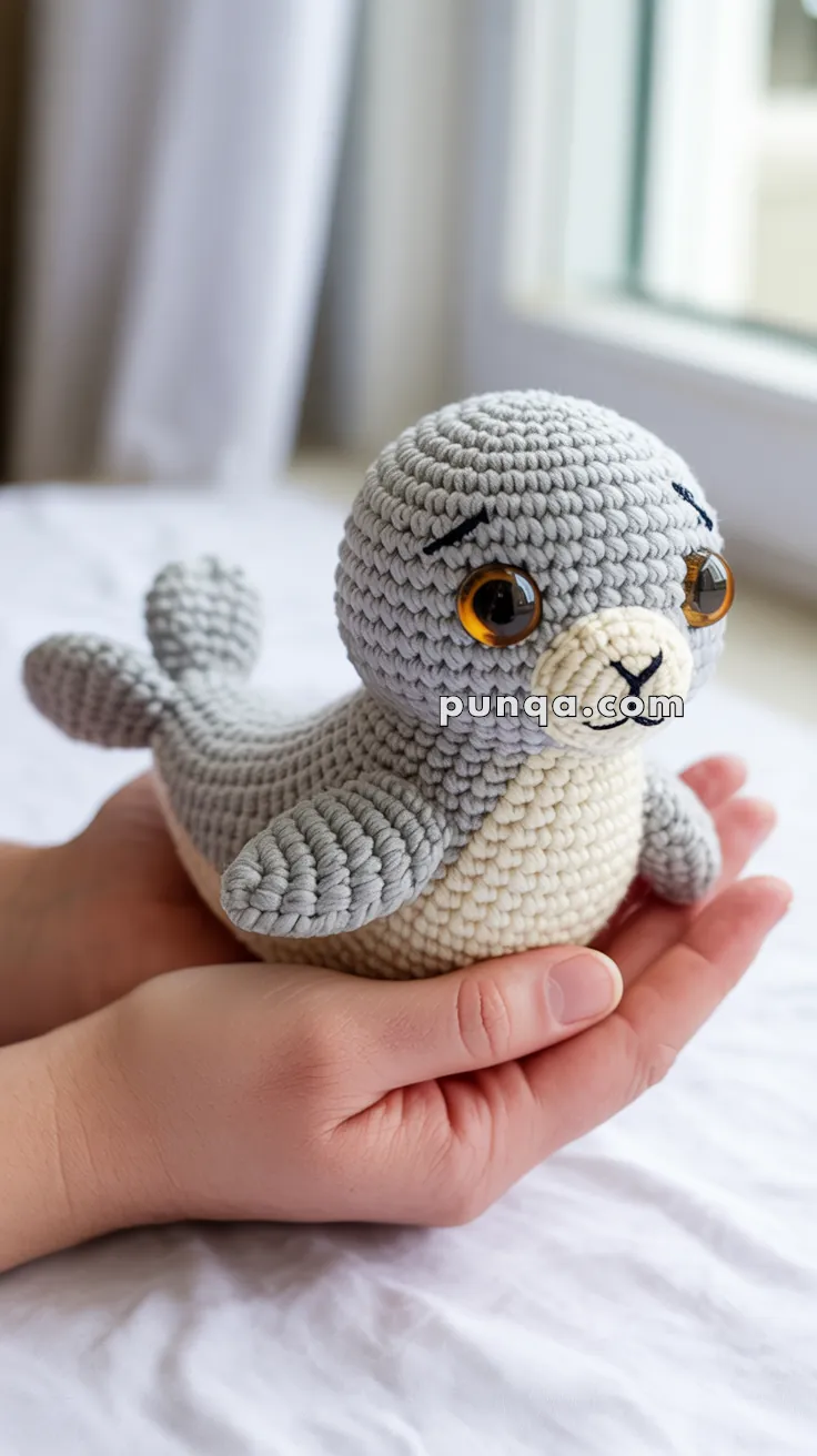 A person holds a small, crocheted seal toy with large amber eyes, featuring gray and cream-colored yarn, on a bed near a window.