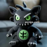 A crocheted plush toy resembling a fierce creature with large green eyes and sharp teeth, sitting on a dark surface. The toy is dark gray with a green emblem on its chest, featuring a black symbol.