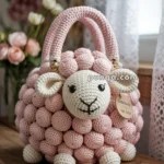 A crocheted handbag shaped like a sheep with a textured pink bobble pattern and a white face, resting on a wooden table. The bag features round, floppy ears and round black eyes, with a tag that reads 'Handmade' attached to it. There are soft-focus flowers and lace curtains in the background.