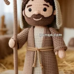A crocheted figure of a shepherd wearing a beige and brown robe, holding a shepherd's crook, standing indoors with straw and festive lights in the background.