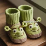 A pair of green knitted slippers featuring a character with a friendly face and ears, placed on a wooden surface next to a window.