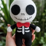 A handmade crochet doll resembling a skeleton, featuring a white head with large black eyes, a stitched smile, and a red bow tie, held in a person's hand with plants in the background.