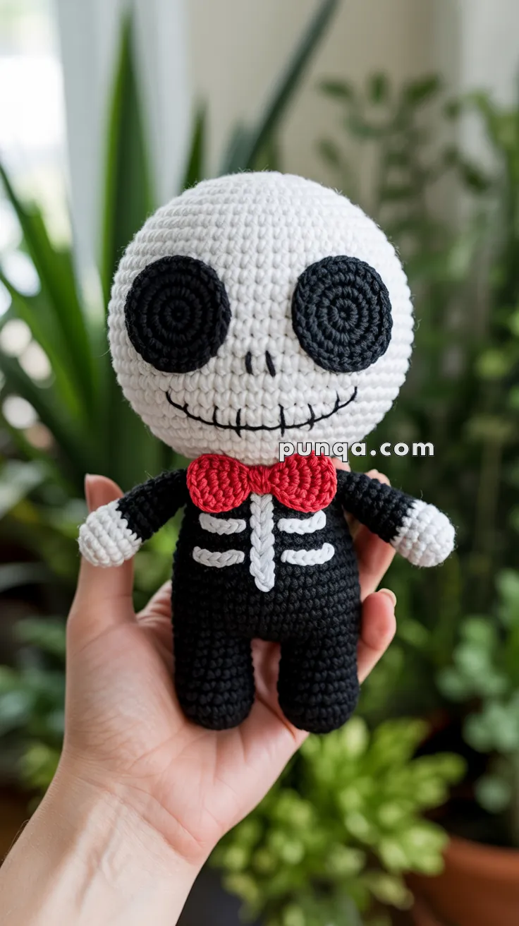 A handmade crochet doll resembling a skeleton, featuring a white head with large black eyes, a stitched smile, and a red bow tie, held in a person's hand with plants in the background.