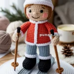 A crochet doll depicting a skier, wearing a red jacket, blue pants, and a white knit hat, holds ski poles while standing on crocheted skis, displayed on a white circular mat.