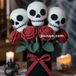 A crocheted bouquet featuring three skulls and red roses, arranged in a black vase with a red bow, set on a wooden table with cobweb decorations and lit candles in the background.