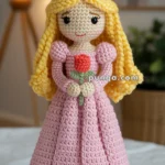 A crocheted doll with long blonde hair, wearing a golden crown and a pink dress, holding a red rose with green leaves.