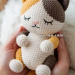 A crocheted stuffed toy cat with closed eyes, featuring white, brown, and orange yarn, being held gently in hands.