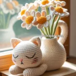 A crocheted white cat figure with closed eyes rests atop a book labeled 'LOOKBOOK,' beside a crocheted bouquet of daisies in a jug on a sunlit windowsill.