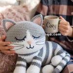 A person in plaid pajamas holds a steaming mug labeled "Cozy" while sitting next to a plush, crocheted gray and white striped cat on a sofa.