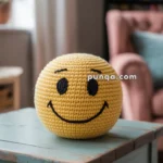 A crocheted yellow smiley face ball sits on a rustic teal wooden table, with a cozy room featuring a pink armchair and bookshelves in the background.