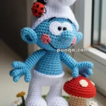 A crocheted blue character wearing a white hat and pants, with a red ladybug on the hat, stands next to a red and brown mushroom on a green base with white flowers.