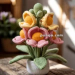 A crochet arrangement of colorful snapdragon flowers with yellow centers and pink and yellow petals, placed in a small white pot on a wooden surface.