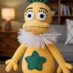 A crocheted yellow toy resembling a cartoon character with big eyes, a round nose, and green hair-like protrusions. It features a white ruffled collar and a green star on its chest, sitting on a wooden surface in a cozy room with bookshelves in the background.
