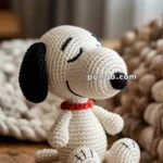 A crocheted toy dog with a red collar, black ears, and a smiling expression, sitting on a wooden surface.
