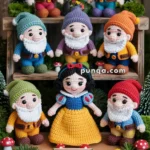 Crocheted figures of Snow White and the seven dwarfs are arranged on a wooden display, surrounded by small plants and mushrooms, with a sign reading 'Snow White's Forest.' Each dwarf has a colorful outfit and white beard, while Snow White wears a blue and yellow dress with a red bow in her hair.