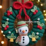 A festive crochet Christmas wreath featuring a snowman with a green and red hat and scarf, surrounded by red berries and green leaves, and topped with a large red bow. The wreath is hanging on a wooden door illuminated by warm lights in the background.