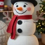 A crocheted snowman with a black hat decorated with holly leaves and berries, a red scarf, and black buttons stands indoors, surrounded by holiday decorations.