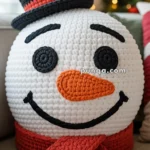 A crocheted snowman pillow with a black top hat, an orange carrot-like nose, and a red scarf, sitting on a couch with a blurred Christmas tree in the background.