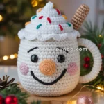 A crocheted mug with a smiling face, topped with white whipped cream and colorful sprinkles, and a candy-like handle, set in a festive holiday scene with pine branches, baubles, and warm lights in the background.