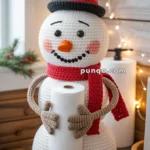 A crocheted snowman toilet paper holder with a black top hat, red scarf, and carrot nose, holding a roll of toilet paper, set in a festive decor with Christmas lights and greenery.