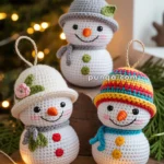 Three crocheted snowmen ornaments with colorful hats and scarves, displayed on a wooden surface with Christmas lights and greenery in the background.