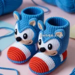 Crocheted baby booties styled to resemble a cartoon character, featuring blue yarn with red, white, and tan accents, surrounded by balls of blue yarn on a pastel background.