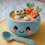 A crocheted bowl of ramen with a smiling face, garnished with crocheted carrots and tofu, placed on a yellow crocheted mat with a matching spoon.