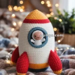A crochet toy rocket with red and yellow accents, featuring a smiling astronaut in the window, set against a blurred background with soft, glowing lights.