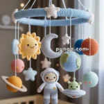 A crocheted baby mobile with space-themed figures such as a sun, moon, planets, an astronaut, and an alien in a spaceship, hanging above a crib.