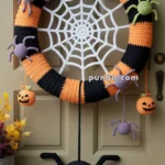 A crocheted Halloween wreath on a wooden door, featuring orange and black stripes with attached spiders and jack-o'-lanterns. A white crochet spider web is at the center, with a hanging black spider below. Decorative leaves and a metal bucket with foliage sit on the doorstep.