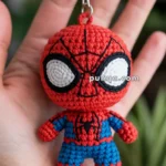 A handmade crochet keychain featuring a small figure in red and blue resembling a well-known superhero, held in the palm of a hand.