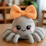 A crocheted plush toy resembling an octopus with a large orange bow on its head, featuring a cute face with rosy cheeks and black eyes, placed on a wooden table in a cozy room setting.