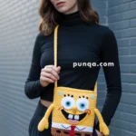 A woman wearing a black turtleneck and pants is holding a crochet handbag designed to resemble SpongeBob SquarePants, standing against a gray brick wall.