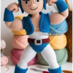 A crocheted doll wearing a blue helmet, goggles, and a sleeveless blue vest, with white pants and blue boots, standing in front of a background with pastel yarn balls and crochet hooks.