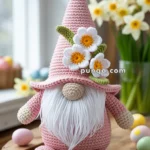 A knitted gnome with a pink hat adorned with white flowers and a green leaf sits on a wooden table, with daffodils and colorful Easter eggs in the background.