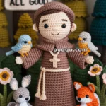 Crocheted doll of a friar, wearing a brown robe, holding birds on each arm, surrounded by crocheted animals including a rabbit and a fox, with a sign in the background reading "Peace and All Good."