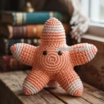 A crocheted orange and white starfish toy with black eyes, placed on a wooden surface against a backdrop of stacked books and a curtained window.
