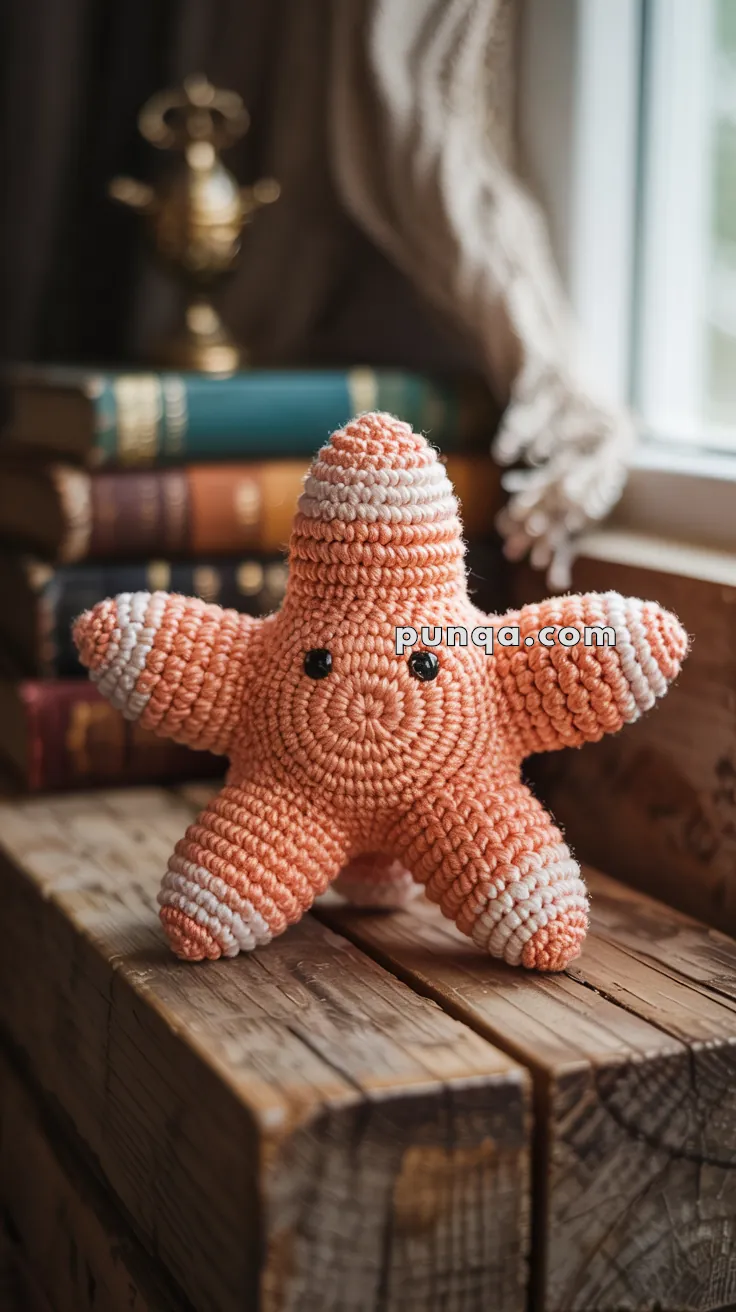 A crocheted orange and white starfish toy with black eyes, placed on a wooden surface against a backdrop of stacked books and a curtained window.